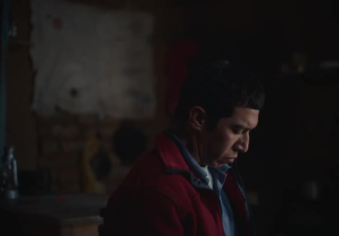 A man in a red jacket looks down thoughtfully in a dimly lit workshop with a cluttered background