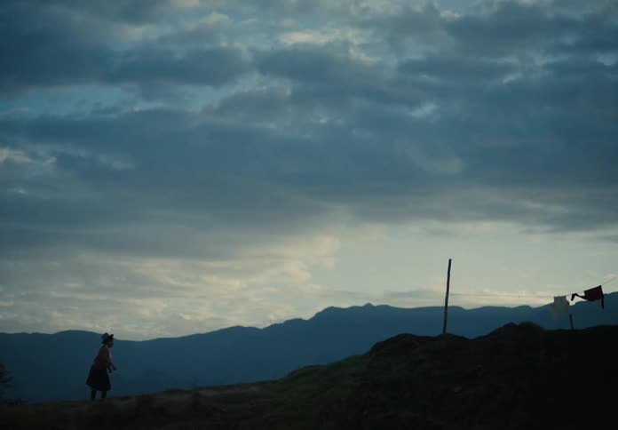 A woman runs toward a small brick house on a quiet hillside at dusk
