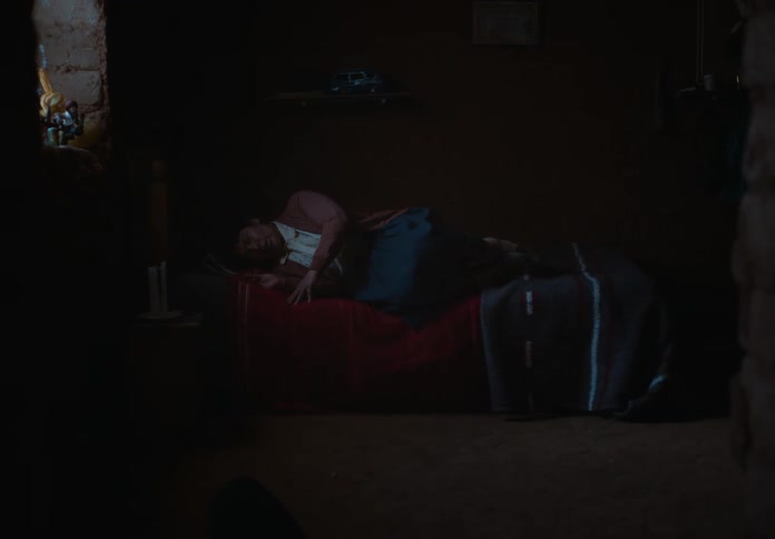 A person sits in a dimly lit room appearing to be in a state of distress the person is sitting on a bed and looking down at their hands