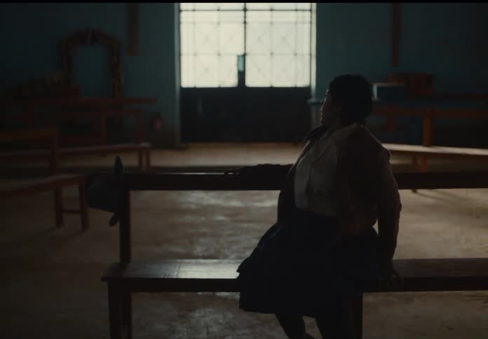 A woman previously lying down on a wooden church pew sits up and looks towards a large gridded window at the front of the dark church