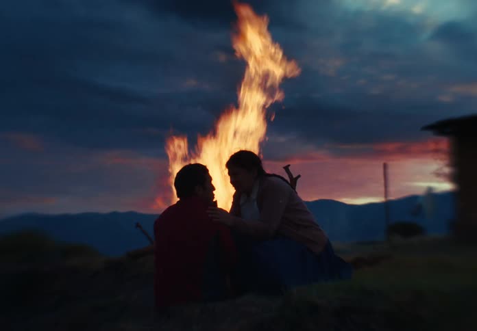 A man sits near a blazing campfire at dusk talking to a person silhouetted against a dramatic mountain sunset