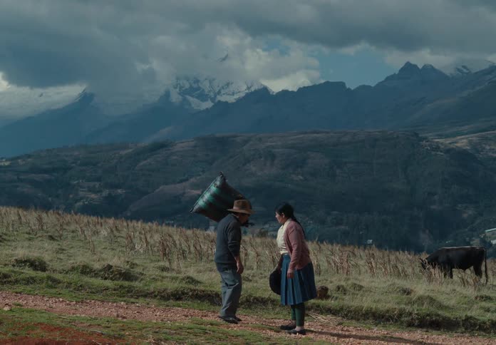 A man walks along a dirt path carrying a heavy sack as a woman approaches snowcapped mountains looming in the distance