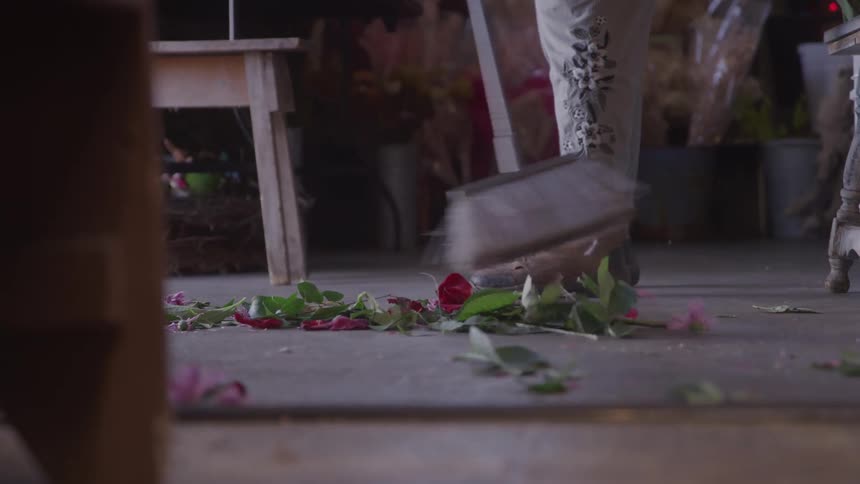 A person in a flower shop sweeps a pile of discarded rose petals and leaves from the concrete floor with a broom
