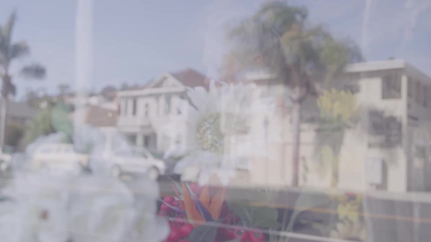A woman with blonde hair arranges flowers in the window of a flower shop as the blurred reflection of a neighborhood street with buildings and cars shimmers across the glass