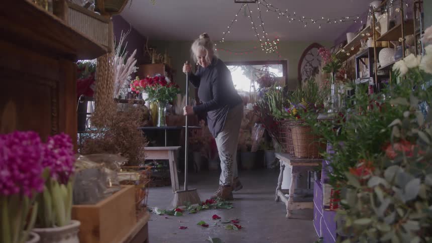 A female florist sweeps discarded petals from the floor of her charmingly cluttered shop which is filled with various floral arrangements and decorated with string lights