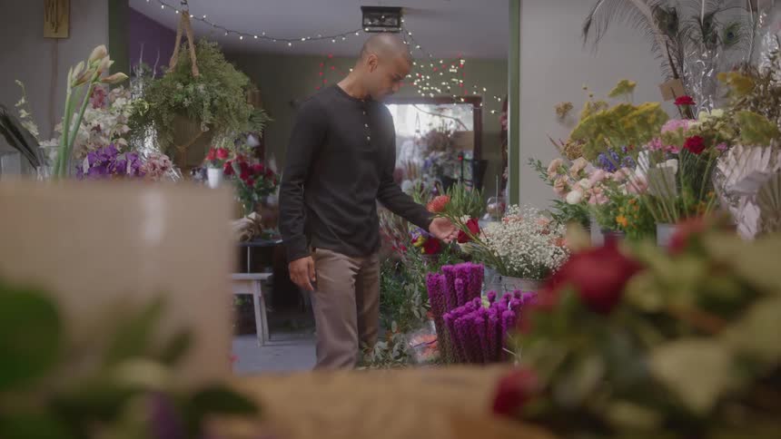A man walks through a flower shop surrounded by colorful floral arrangements and soft string lights