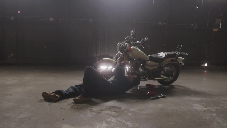 Man lying on the ground performs maintenance on a motorcycle inside a dimly lit warehouse