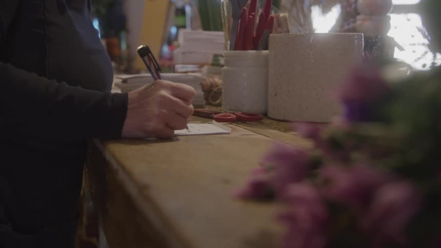 A woman writes on a piece of paper on a shelf with a flower plant beside her and a mirror on the wall