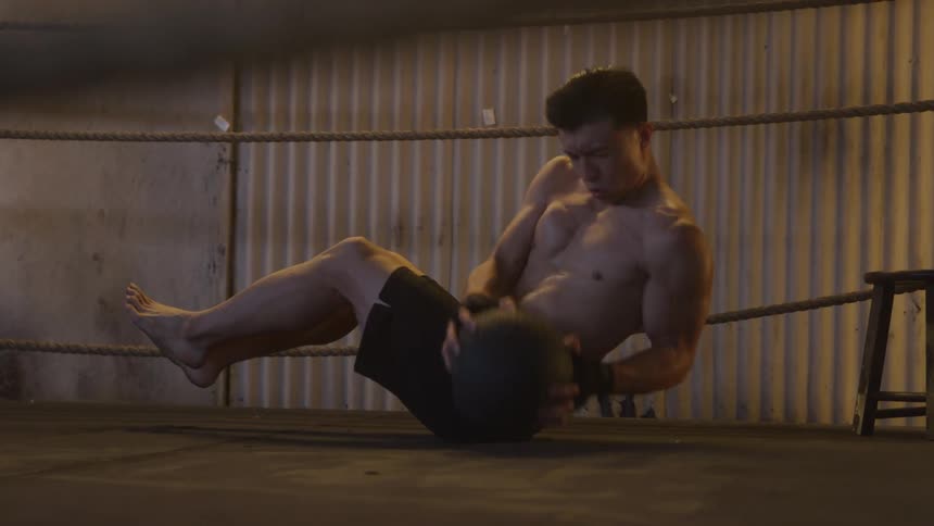 A young man wearing trunks and shirtless works out indoors in a gym doing sidetoside movements while holding a ball