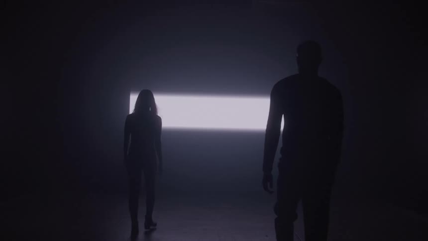 Two silhouettes walk toward an intense strip of light in the dark room