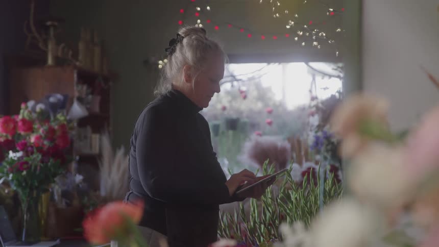 Blonde woman interacts with a tablet among flowers inside a flower shop decorated with lights