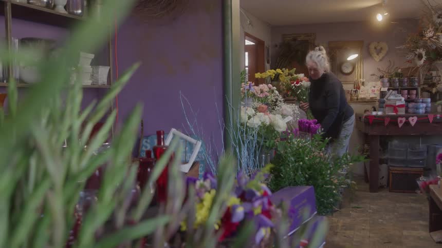 A female florist with blonde hair in a bun arranging a vibrant bouquet of flowers inside of a brightly lit flower shop