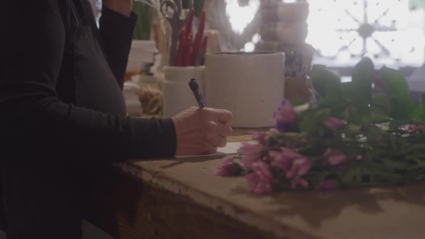 Woman writing on a piece of paper beside pink flowers on a flower shop counter