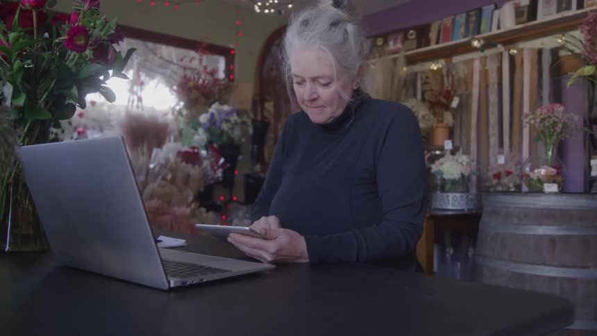 An elderly woman with gray hair sits behind a desk with a computer on top using her mobile phone with a flower plant on the side of the desk