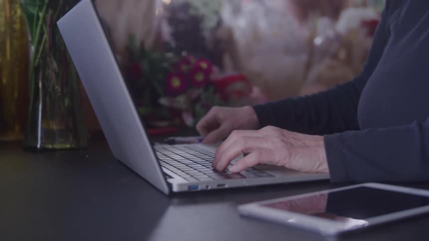 A female business owner is sitting at a wooden desk with a tablet and a bouquet of flowers next to her typing on a laptop computer and writing on a notepad