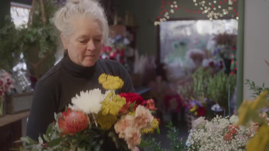 A female florist with her blonde hair pulled into a messy bun is holding a large beautiful bouquet of red yellow and white flowers in a cluttered flower shop