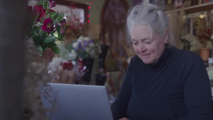 Woman smiles while working on a laptop surrounded by flowers in her shop