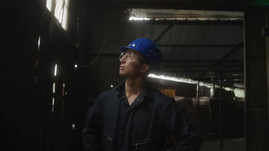 A man wearing a blue hard hat and safety glasses stands in a dimly lit industrial building looking upward thoughtfully