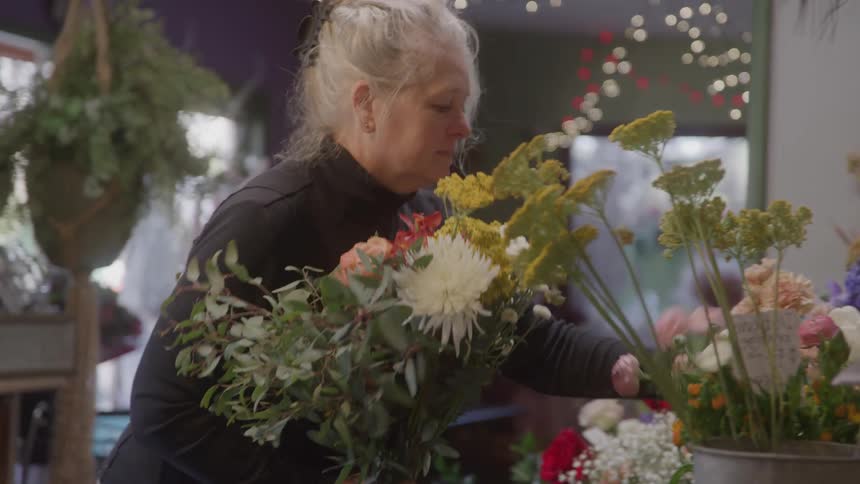 A female florist with blonde hair pulled back holds a large bouquet of white and yellow flowers with green foliage as she works in her brightly lit cluttered flower shop