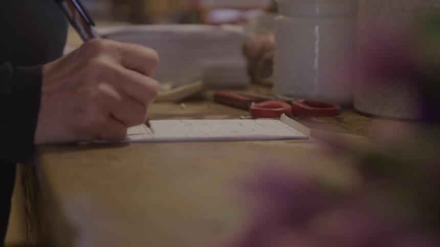A person writes on a notepad at a rustic wooden table surrounded by jars scissors and blurred flowers in the foreground