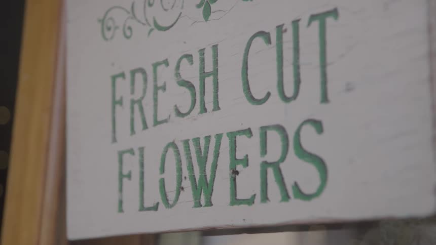 A white wooden sign with faded green lettering that reads fresh cut flowers is hanging next to a window