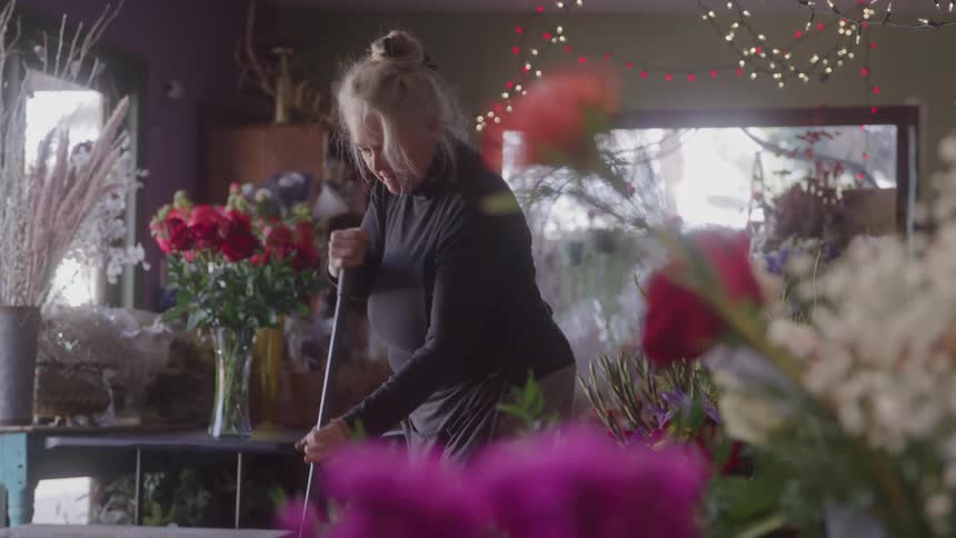 A blonde female florist in a black turtleneck and apron sweeps a wooden floor in a flower shop with a longhandled broom