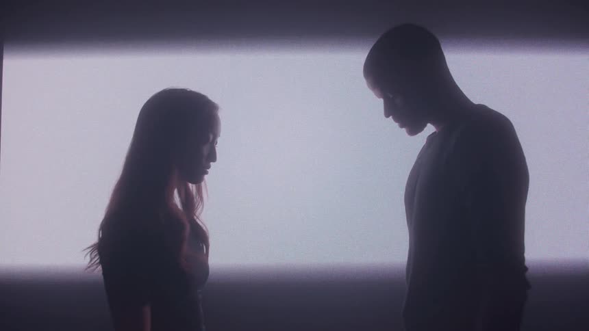 A silhouetted man and woman stand facing each other looking down in front of a brightly lit wall that is bordered by two horizontal expanding dark stripes