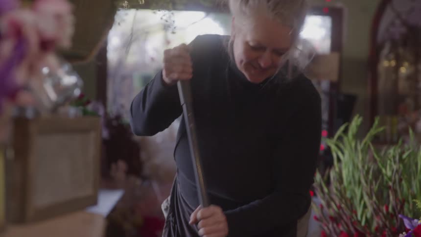 Older woman cleans the floor of a flower shop surrounded by colorful arrangements and plants