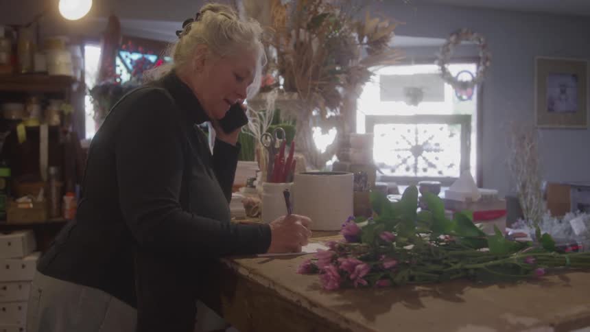 A female florist with blonde hair in a messy bun stands behind a wooden counter in her shop talking on a smartphone while writing on a piece of paper next to a small pile of pink flowers