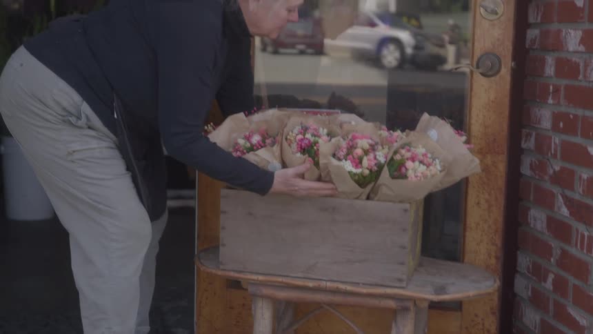 A woman holds a bouquet of flowers and a wooden box walking out of a shop with an open sign on the door the sunlight is bright creating a lens flare as she exits the building