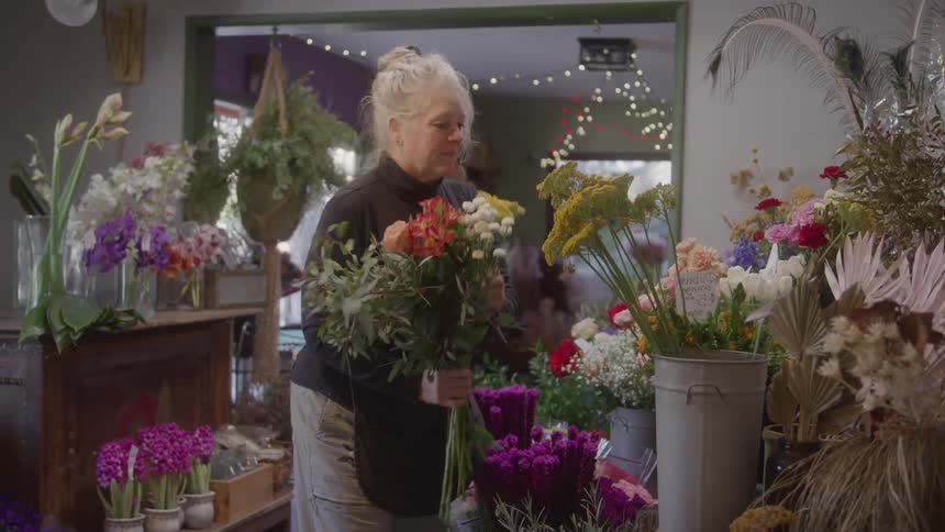 Woman holding a bouquet of mixed flowers while arranging displays in a colorful flower shop