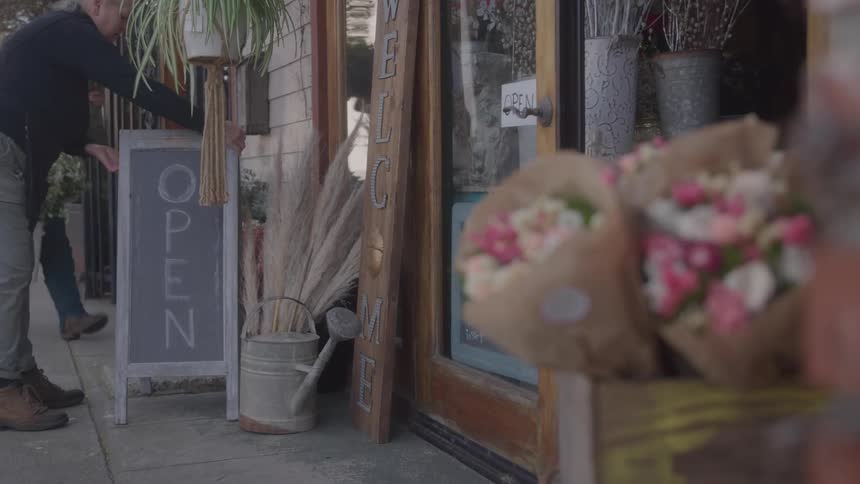 A man puts an open sign in front of a flower shop adorned with welcome signs and fresh bouquets