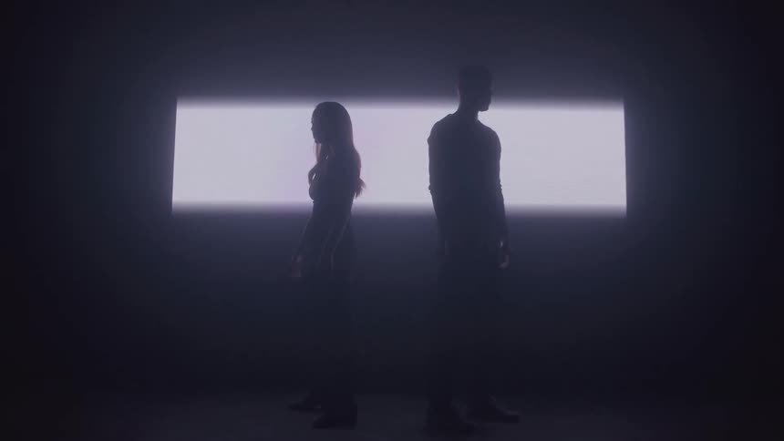 A man and a woman in silhouette are standing apart from each other in a dark room in front of a bright white rectangular light on the back