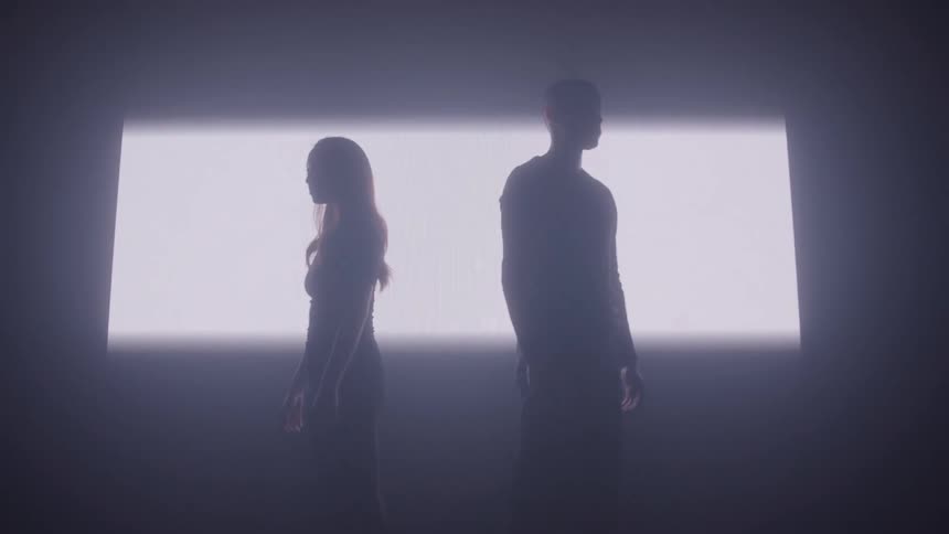 A man and a woman stand in front of a large screen in a dark room with the screen briefly illuminating the room before going dark