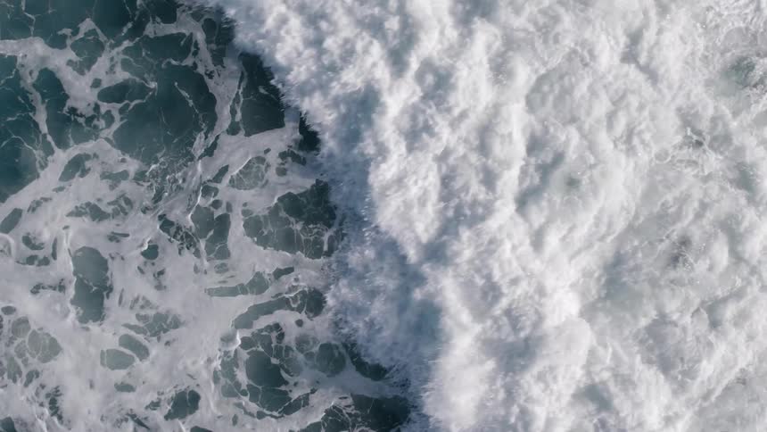 A topdown aerial view shows a large ocean wave crashing and filling the screen with turbulent white seafoam