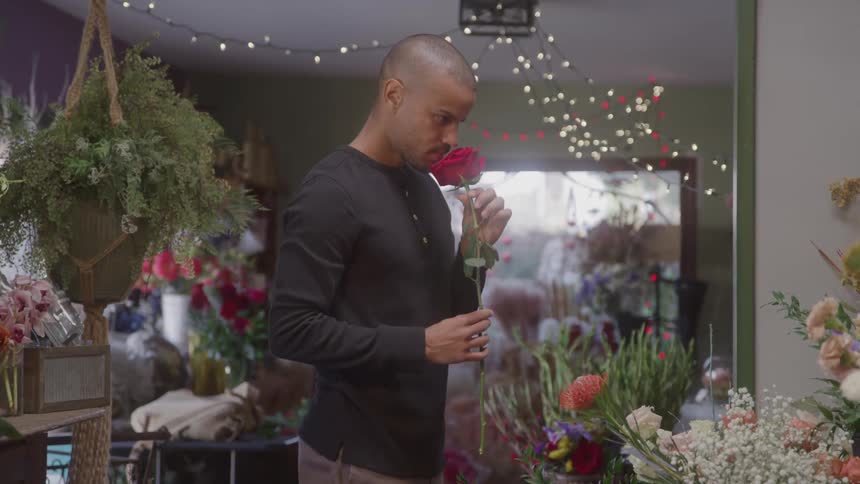 A man inside a flower shop picks up a rose and smells it at midday