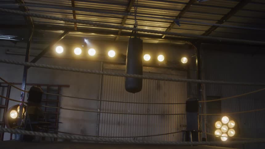 A boxing ring with ropes is shown in an industriallooking space with a large black punching bag hanging in the center the area is illuminated by several round bright lights mounted overhead on the metal ceiling