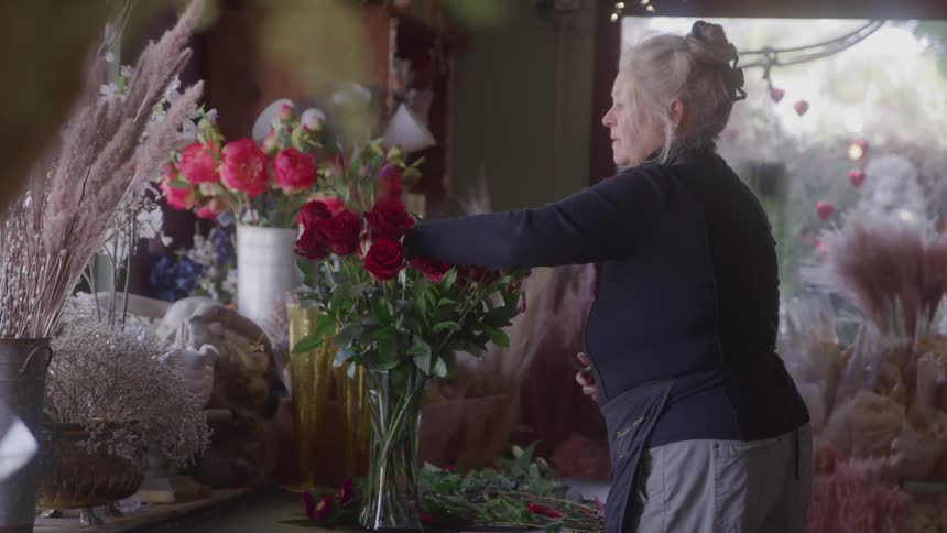 A woman arranges red roses in a vase inside a flowerfilled shop softly lit by natural light