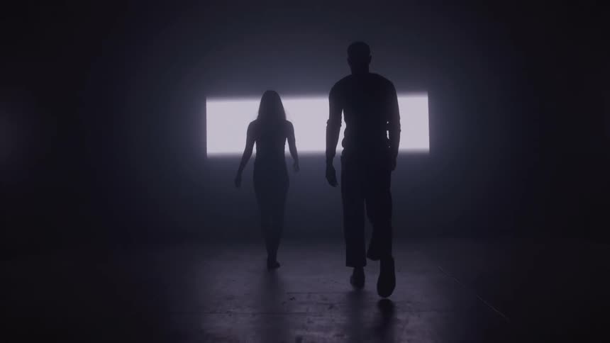 A man and a woman seen in silhouette walk toward the camera from a bright rectangular light source in the background of a dark room