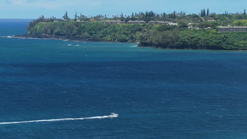 A white speedboat travels across the deep blue ocean leaving a wake behind it near a lush tropical coastline with buildings