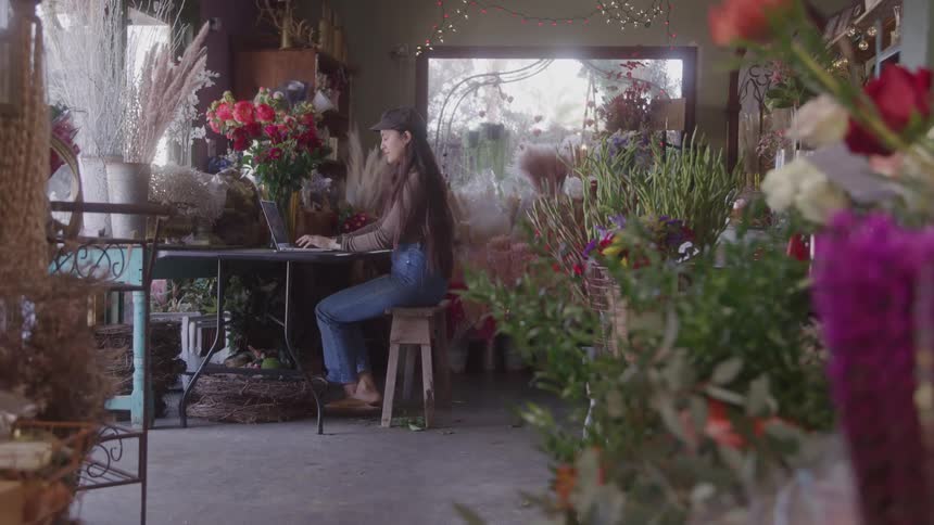 Woman sitting on a stool working on a laptop surrounded by floral arrangements inside a flower shop