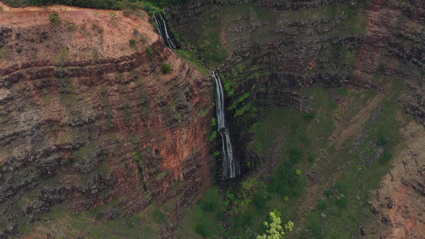 A thin waterfall cascades down the immense rugged cliffs of a canyon revealing layers of red earth and green vegetation