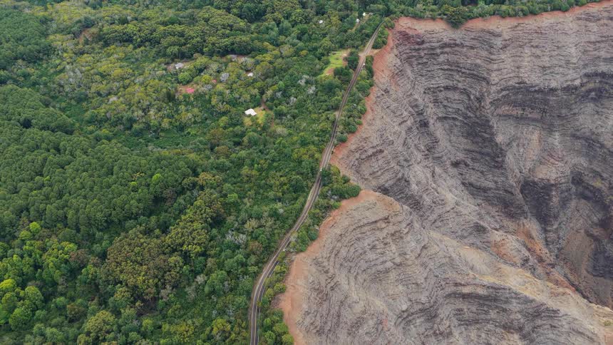 An aerial view shows a sharp contrast between lush green forest and a vast layered rocky cliff divided by a winding road