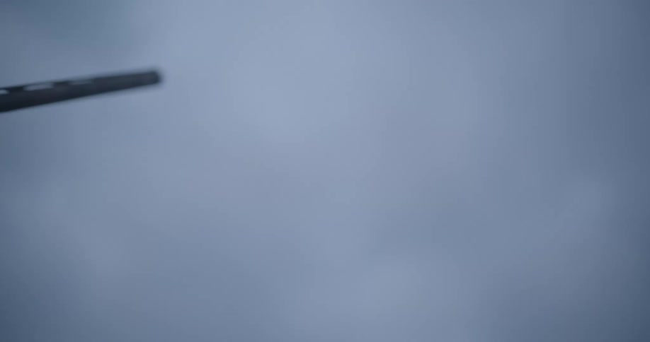 A shotgun barrel angles upward silhouetted against a hazy gray sky as it fires