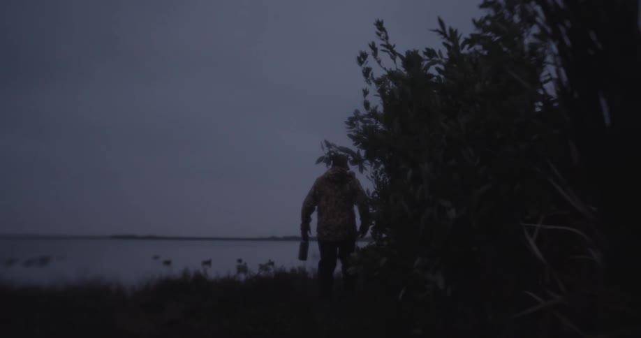 Man walking near a lake at dusk partially obscured by bushes