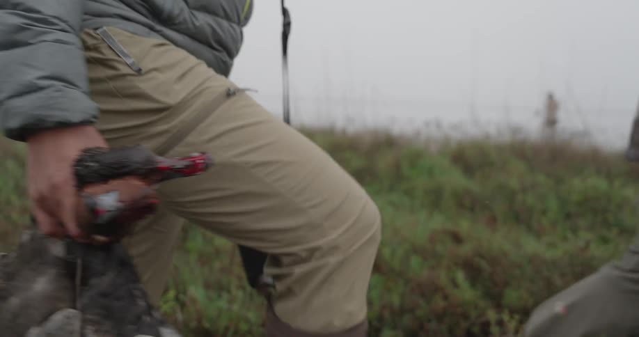 A male hunter wearing a gray puffer jacket and tan waders holds a shotgun and a pair of hunted ducks by their necks while walking through a field on a foggy morning
