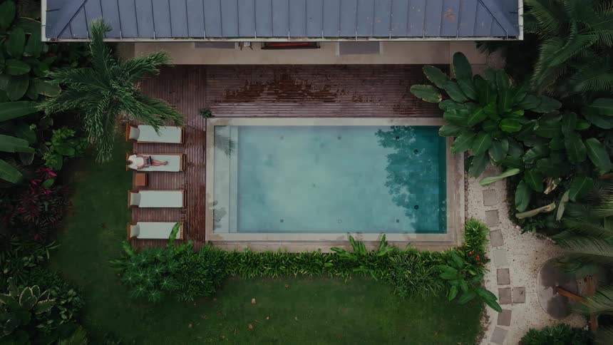 A man sits on a pool chair in an outdoor pool area in the backyard of a building surrounded by trees at midday