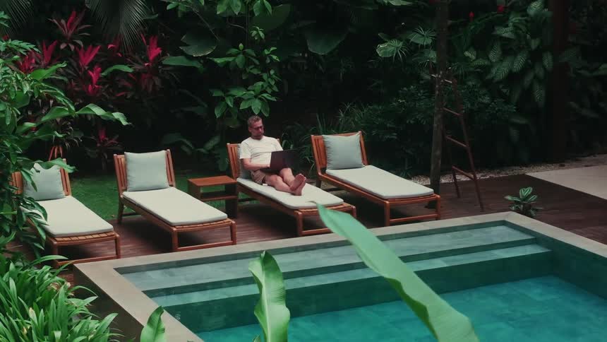 A man lounges on a poolside chair with a laptop surrounded by lush tropical greenery