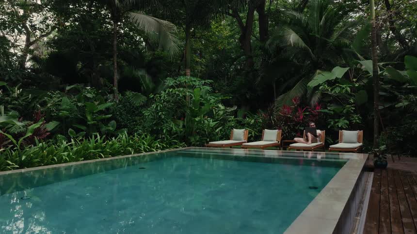 Woman sitting on a lounge chair beside a pool surrounded by dense tropical vegetation
