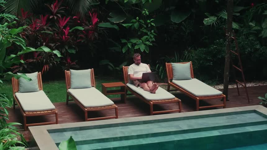 Man sitting on a lounge chair works on a laptop beside a pool surrounded by tropical plants